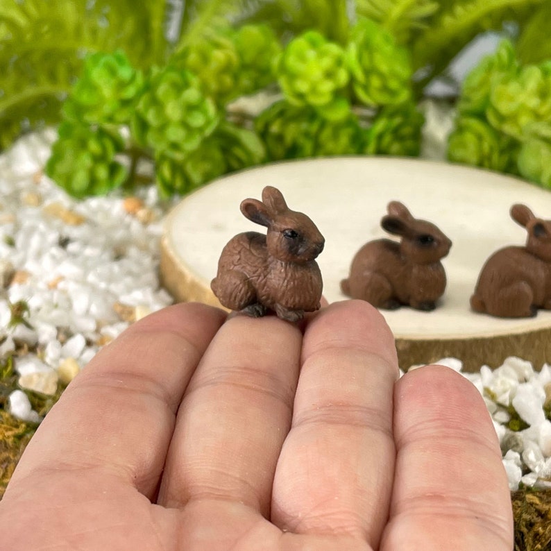 May include: Miniature brown bunny figurines. The small, detailed rabbits are in a sitting position, with their ears perked up. The bunnies are a dark brown color. The background includes greenery and a wooden slice.