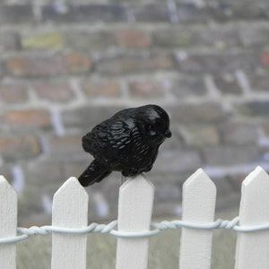 May include: A glossy black bird figurine perched on a white picket fence with a twisted wire detail. The bird is facing left, with a brick wall in the background. The fence has pointed tops.