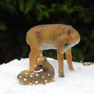 May include: A brown and white deer figurine standing on a white surface with a smaller brown and white deer figurine lying down in front of it.