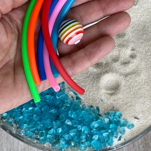 May include: A hand holding a rainbow of colourful rubber tubing with a striped ball in the centre. The tubing is arranged in a semi-circle shape. The background is a glass bowl filled with blue glass beads and white sand.