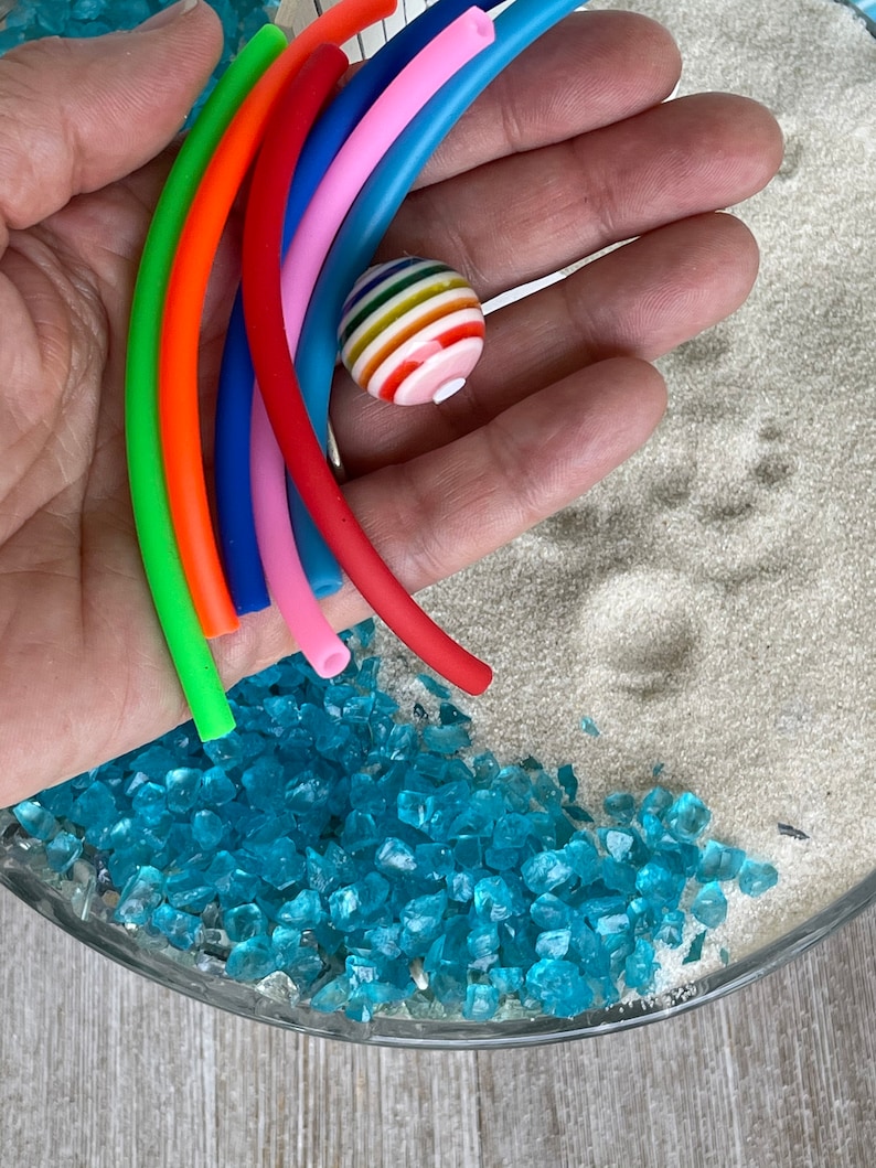 May include: A hand holding a rainbow of colourful rubber tubing with a striped ball in the centre. The tubing is arranged in a semi-circle shape. The background is a glass bowl filled with blue glass beads and white sand.
