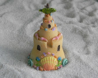 May include: A small sandcastle ornament in a light tan hue, adorned with pink crab claws, blue and yellow seashells, and a green leaf. The castle has tiny windows and is placed on white sand, evoking a beach scene.