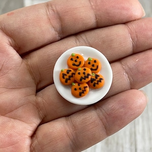May include: A small white plate holds six miniature orange pumpkin-shaped cookies with black facial features and green stems. The cookies are arranged in a circular pattern, perfect for Halloween-themed crafts or decorations.