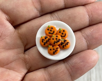 May include: A small white plate holds six miniature orange pumpkin-shaped cookies with black facial features and green stems. The cookies are arranged in a circular pattern, perfect for Halloween-themed crafts or decorations.