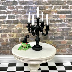 May include: A black metal candelabra with five white candles sits on a white pedestal table. A green frog wearing a black witch's hat sits on the table next to the candelabra.