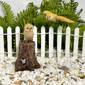 May include: A miniature owl figurine perched on a detailed tree stump, with a small mouse peeking out from a hole. A second owl figurine is in flight. The scene is set against a white picket fence and greenery.