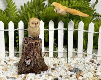 Könnte beinhalten: Eine Miniatur-Eulenfigur sitzt auf einem detaillierten Baumstumpf, mit einer kleinen Maus, die aus einem Loch schaut. Eine zweite Eulenfigur ist im Flug. Die Szene spielt vor einem weißen Zaun und Grün.