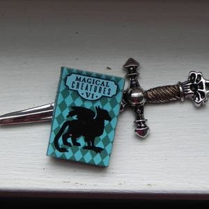 May include: A silver sword with a blue and green book attached to it. The book cover has the text "Magical Creatures - VI."