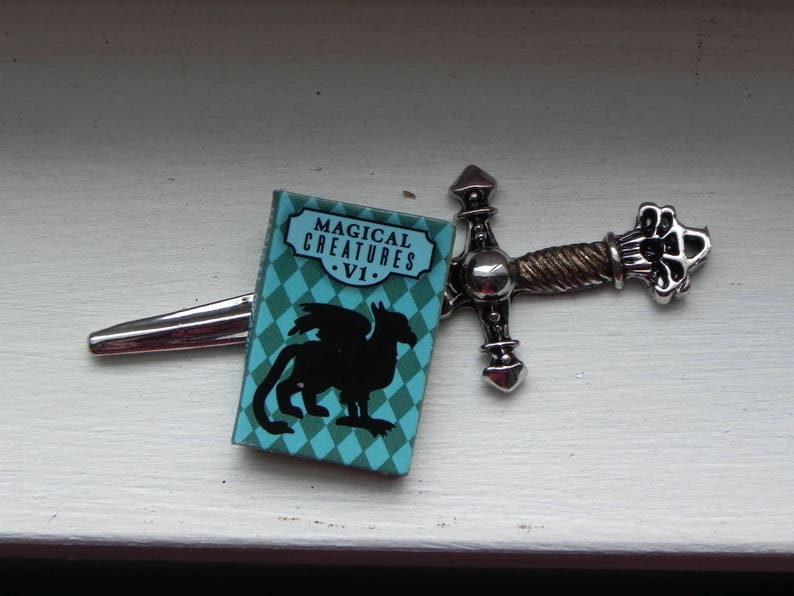 May include: A silver sword with a blue and green book attached to it. The book cover has the text "Magical Creatures - VI."