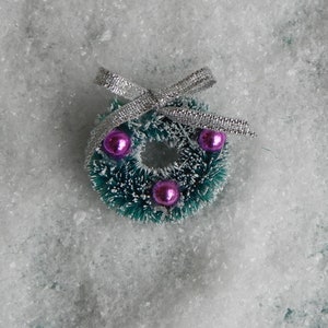 May include: A small green Christmas wreath with a silver bow and three pink ornaments. The wreath is sitting on a white, fluffy surface.