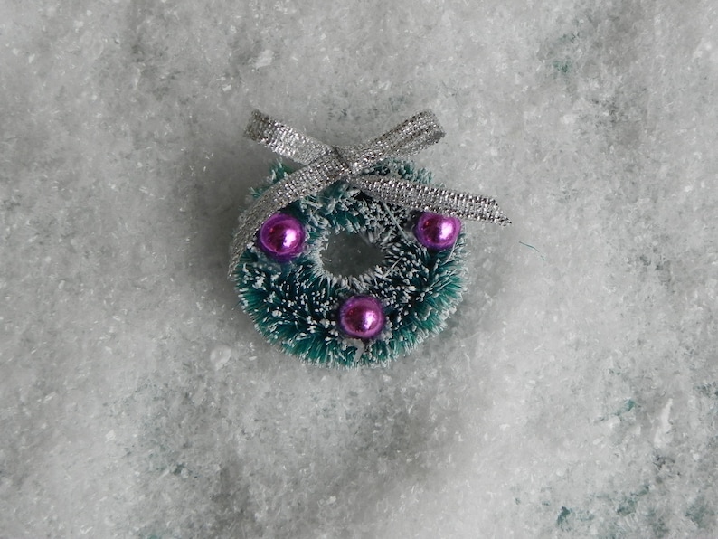 May include: A small green Christmas wreath with a silver bow and three pink ornaments. The wreath is sitting on a white, fluffy surface.