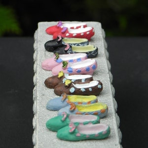 May include: A row of miniature shoes in various colors and patterns, including pink, blue, green, brown, and black, displayed on a white pedestal with decorative trim.