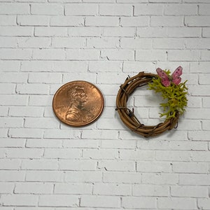 May include: A small, rustic twig wreath with a pink butterfly and green moss accents. The wreath is about the size of a US penny.