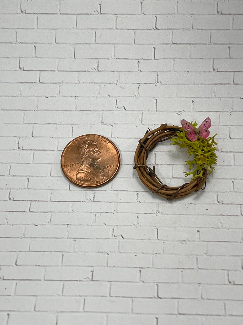 May include: A small, rustic twig wreath with a pink butterfly and green moss accents. The wreath is about the size of a US penny.
