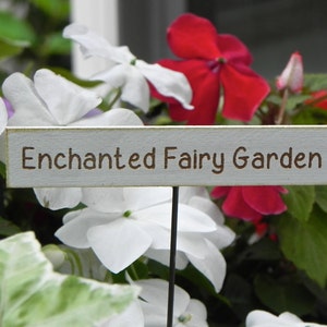 Op de afbeelding: Een houten bord met de tekst "Enchanted Fairy Garden" in bruine letters, tegen een achtergrond van levendige rode, witte en roze bloemen en groen blad. Het bord staat op een zwarte metalen paal.