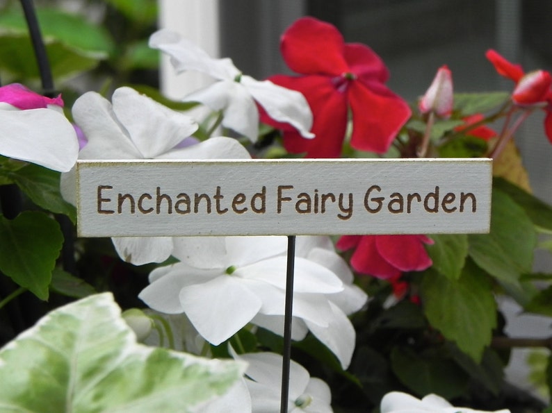 Op de afbeelding: Een houten bord met de tekst "Enchanted Fairy Garden" in bruine letters, tegen een achtergrond van levendige rode, witte en roze bloemen en groen blad. Het bord staat op een zwarte metalen paal.