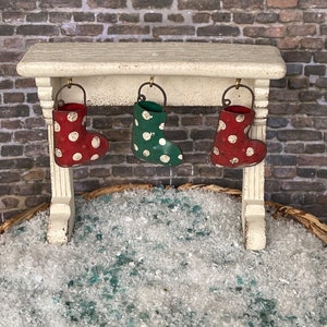 May include: A miniature Christmas scene featuring a white mantel with three small, polka-dotted stockings hanging from it. The stockings are red and green. The scene is set against a brick wall and a bed of faux snow.