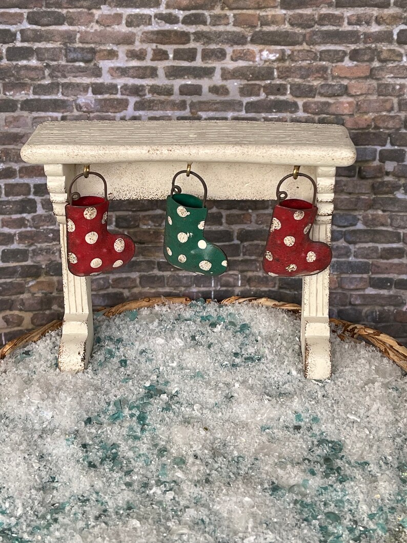 May include: A miniature Christmas scene featuring a white mantel with three small, polka-dotted stockings hanging from it. The stockings are red and green. The scene is set against a brick wall and a bed of faux snow.