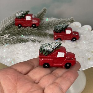 May include: Three red toy pickup trucks with white trim, each carrying a small green Christmas tree with white snow on the branches. The trucks are sitting on a white cake stand with white glitter.