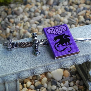May include: A miniature silver sword with a twisted handle and a purple book titled "Magical Creatures - V3." featuring a black dragon illustration.
