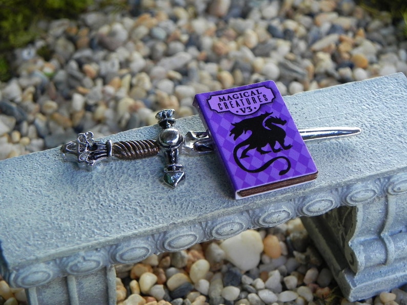 May include: A miniature silver sword with a twisted handle and a purple book titled "Magical Creatures - V3." featuring a black dragon illustration.