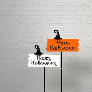 May include: Two Halloween yard signs with black witch hats. One sign is orange with the words "Happy Halloween" in black script. The other sign is white with the same text. Both signs are on black metal stakes.