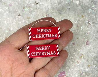 May include: Two small, rectangular Christmas ornaments. They have a red background with the words "MERRY CHRISTMAS" in white. Each ornament features white and red diagonal stripes on the sides and a white wire loop.