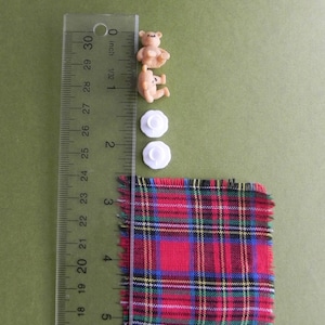 May include: An assortment of craft supplies including two small teddy bear figurines, two white flower-shaped buttons, and a square of red plaid fabric. A ruler shows measurements in inches.