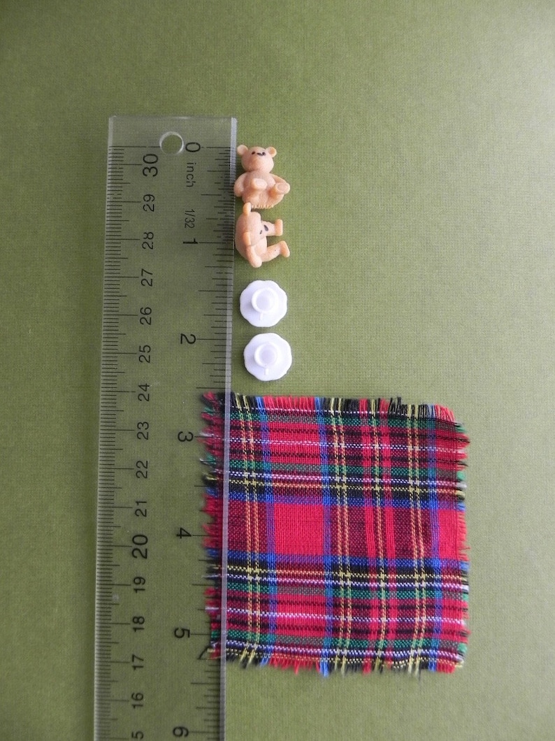 May include: An assortment of craft supplies including two small teddy bear figurines, two white flower-shaped buttons, and a square of red plaid fabric. A ruler shows measurements in inches.
