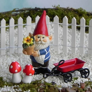 May include: A miniature garden scene featuring a gnome holding a flower pot, a red wagon, and two mushroom figurines. The gnome wears a red hat and blue striped shirt. A white picket fence and small stones complete the scene.