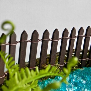 May include: A miniature wooden fence with a brown stain, surrounded by green moss and blue glass stones. The fence is made of small, pointed wooden stakes connected by wire.