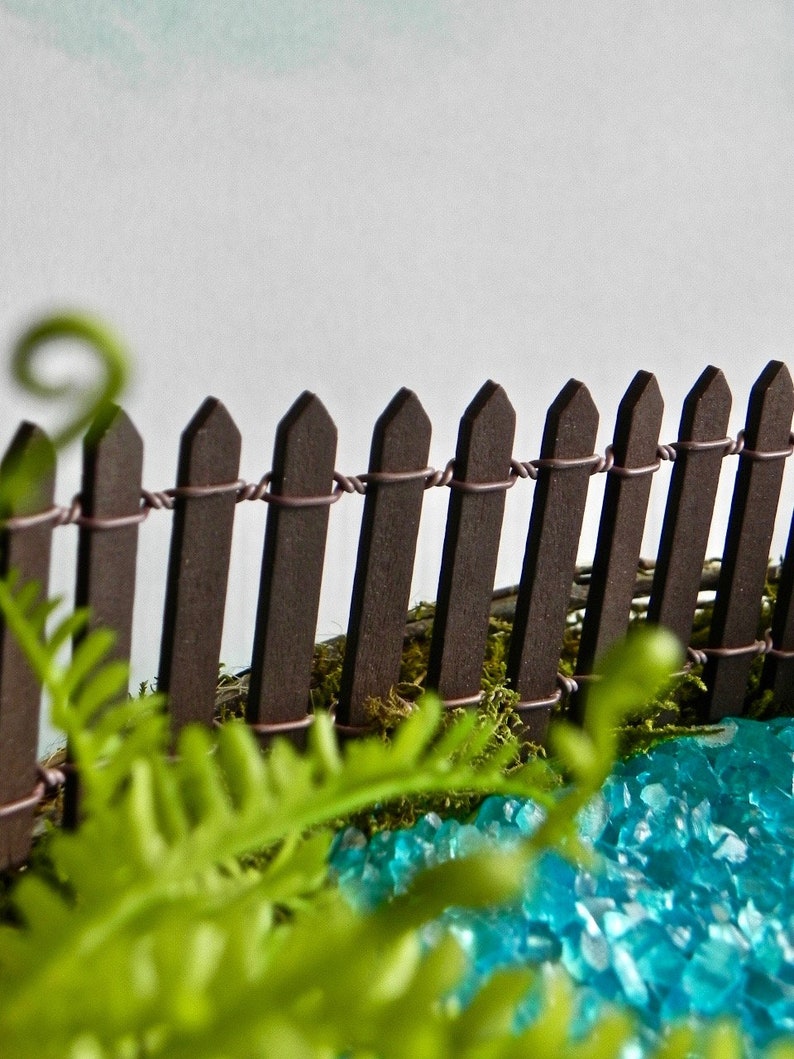 May include: A miniature wooden fence with a brown stain, surrounded by green moss and blue glass stones. The fence is made of small, pointed wooden stakes connected by wire.