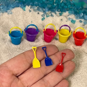 May include: A collection of five miniature plastic buckets in blue, purple, pink, yellow, and red, each with a contrasting handle. Three small plastic shovels in yellow, blue, and red are displayed alongside the buckets in a set.