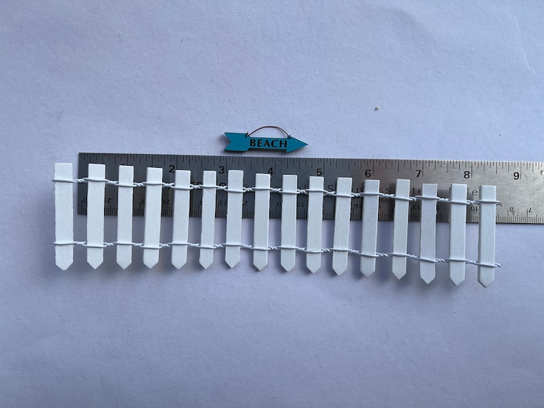 K&ouml;nnte beinhalten: Ein wei&szlig;er Lattenzaun mit einem blauen Holzschild, auf dem "BEACH" steht, das dar&uuml;ber h&auml;ngt. Der Zaun ist etwa 25 cm lang und hat 18 Latten.