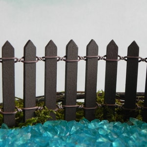 May include: A miniature brown picket fence with wire connecting the top of each picket. The fence is sitting on a bed of turquoise blue glass stones.