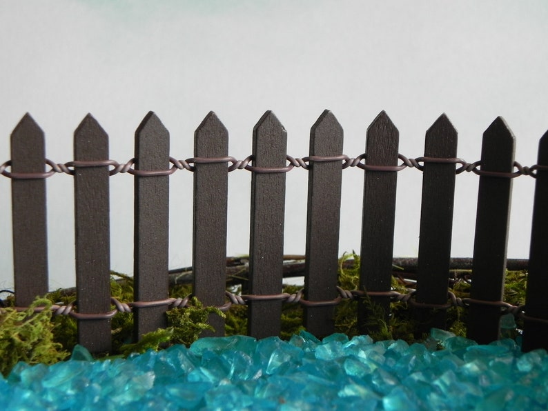 May include: A miniature brown picket fence with wire connecting the top of each picket. The fence is sitting on a bed of turquoise blue glass stones.
