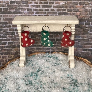 May include: A miniature Christmas scene featuring a white mantelpiece with three hanging stockings. The stockings are red with white polka dots and green with white polka dots. The scene is set against a brick wall and a bed of faux snow.