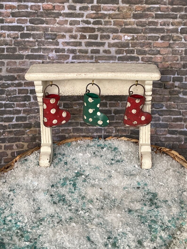 May include: A miniature Christmas scene featuring a white mantelpiece with three hanging stockings. The stockings are red with white polka dots and green with white polka dots. The scene is set against a brick wall and a bed of faux snow.