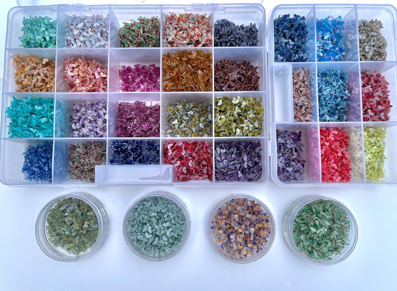 May include: A clear plastic storage box filled with small, colorful, glittery, butterfly-shaped pieces. The box has 24 compartments and is divided into two sections. There are four small jars filled with the same glittery pieces on a white surface.