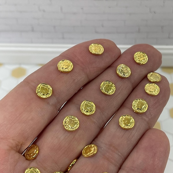 May include: A hand holding a pile of small, gold-colored, round coins with a raised design. The coins are about the size of a dime.