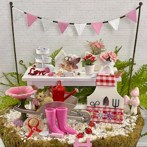 May include: A miniature fairy garden scene with a white table, a red and white gingham apron, a red watering can, a pink flower, a wheelbarrow, and pink rain boots. The table has a sign that says "Love".