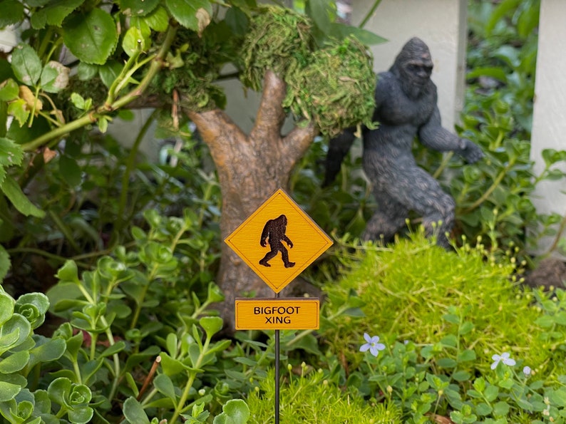 May include: A yellow diamond-shaped sign with a black silhouette of Bigfoot walking, with the text "BIGFOOT XING" on a brown rectangular sign below.