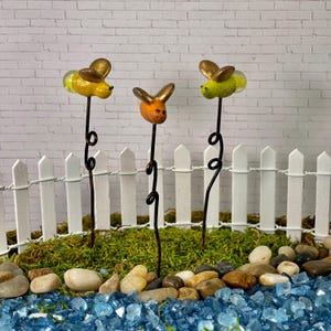 May include: Three miniature bee figurines with gold accents on wire stems. The bees are yellow, orange, and green. The stems are placed in a miniature garden setting with moss, rocks, and blue glass.