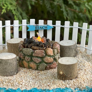 K&ouml;nnte beinhalten: Ein Miniatur-Steinkamin mit einem unechten Feuer und einem blauen Schild mit der Aufschrift "Beach" vor einem wei&szlig;en Lattenzaun. Es gibt vier Miniatur-Holzscheite, die den Kamin umgeben.