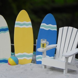 May include: A white Adirondack chair with a blue surfboard and two other surfboards, one yellow and one white, with blue and white stripes. A small beach ball and a glass of beer are on a table in front of the chair.