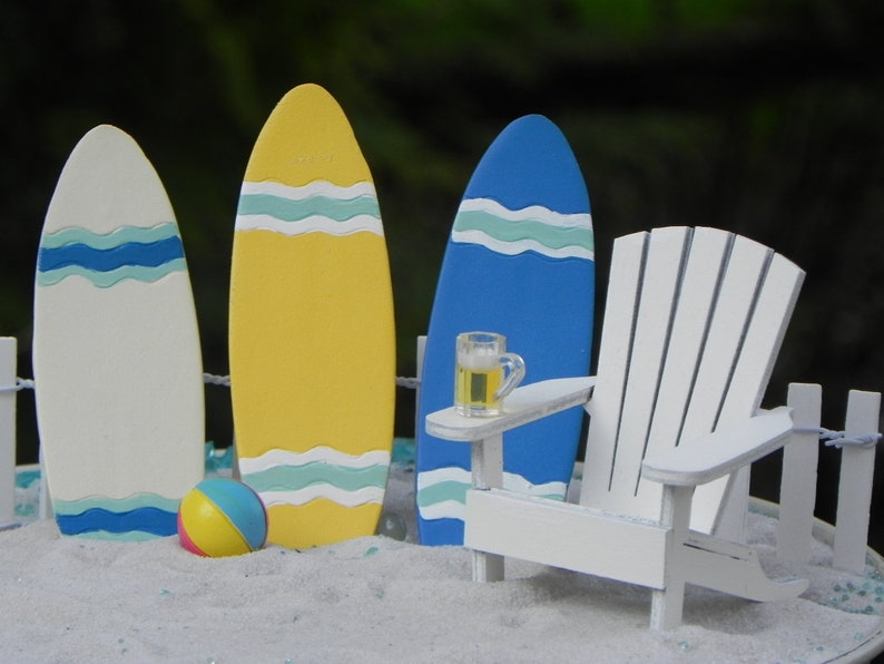 May include: A white Adirondack chair with a blue surfboard and two other surfboards, one yellow and one white, with blue and white stripes. A small beach ball and a glass of beer are on a table in front of the chair.
