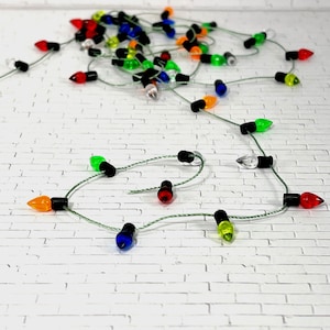 May include: A string of miniature decorative holiday lights featuring small, translucent, multi-colored bulbs in red, green, blue, orange, and clear, each attached to a black base and connected by a thin, light-colored wire.