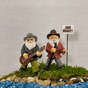 May include: Two figurines of elderly men, one playing a brown guitar and the other holding a microphone. They are dressed in hats, jackets, and pants. A sign reads "JAM NIGHT!" The figurines stand on a bed of moss, pebbles, and blue glass.