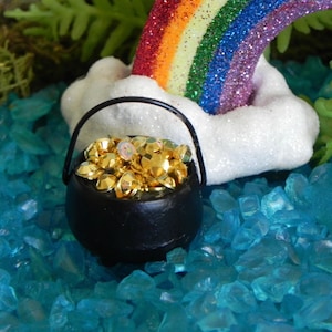 May include: A black pot filled with gold-colored gems sits in front of a white cloud and a rainbow. The pot has a black handle. The background is filled with blue, crystal-like stones, and green foliage.