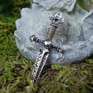 May include: A silver and copper sword pendant with intricate details. The sword has a pointed tip, a cross guard, and a twisted copper handle. The pendant is resting on a grey stone surface.